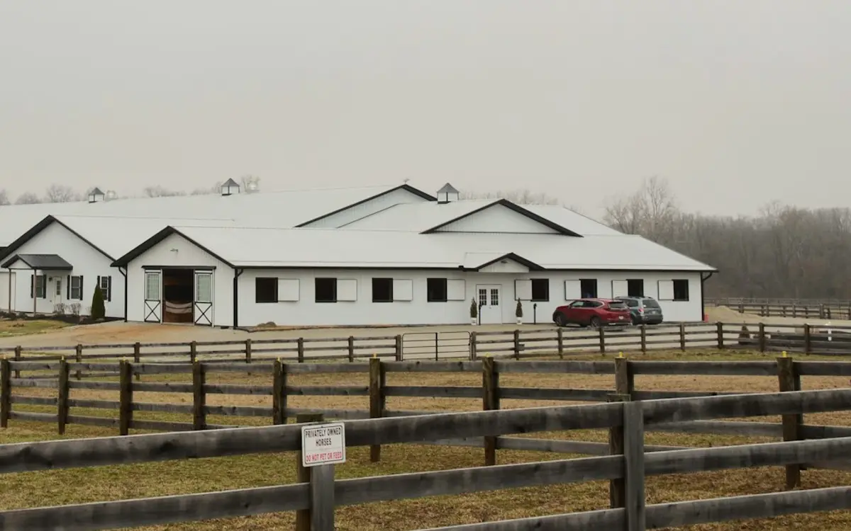 White metal-sided horse barn with cupolas behind board-fenced paddocks
